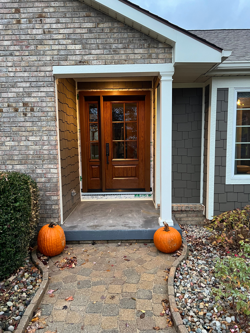 Full front view of a newly installed oak entry door framed by brick siding, pumpkins, and a landscaped path, completed by The Exterior Zone in Lapeer Michigan.